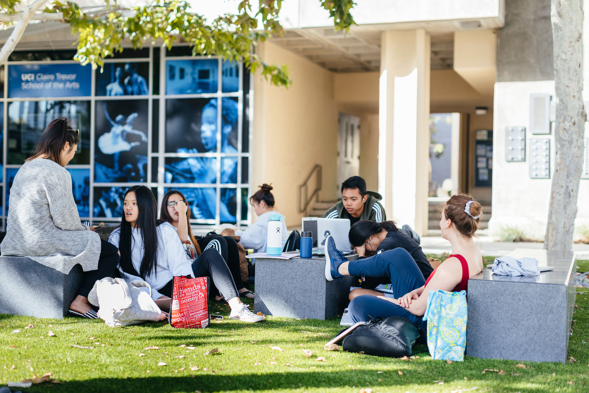 Students relax and study outdoors on campus, sitting on benches and grass with laptops and notebooks near the Claire Trevor School of the Arts building. (Zoomed Out).