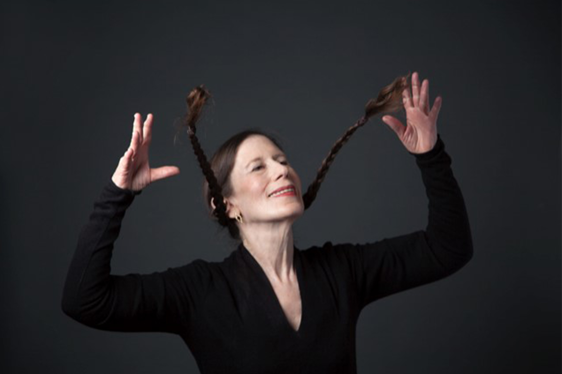 A performer poses expressively against a dark background, raising her hands as her braided hair extends outward.