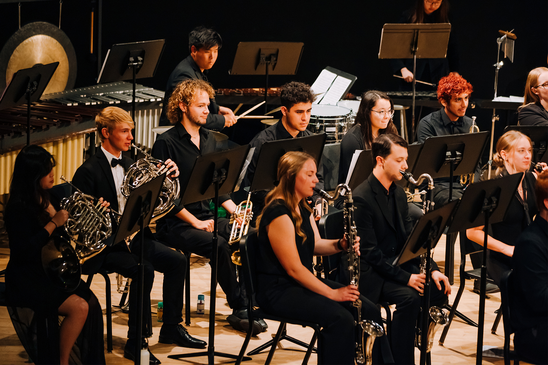 Student wind and brass musicians perform together in a concert ensemble on stage.
