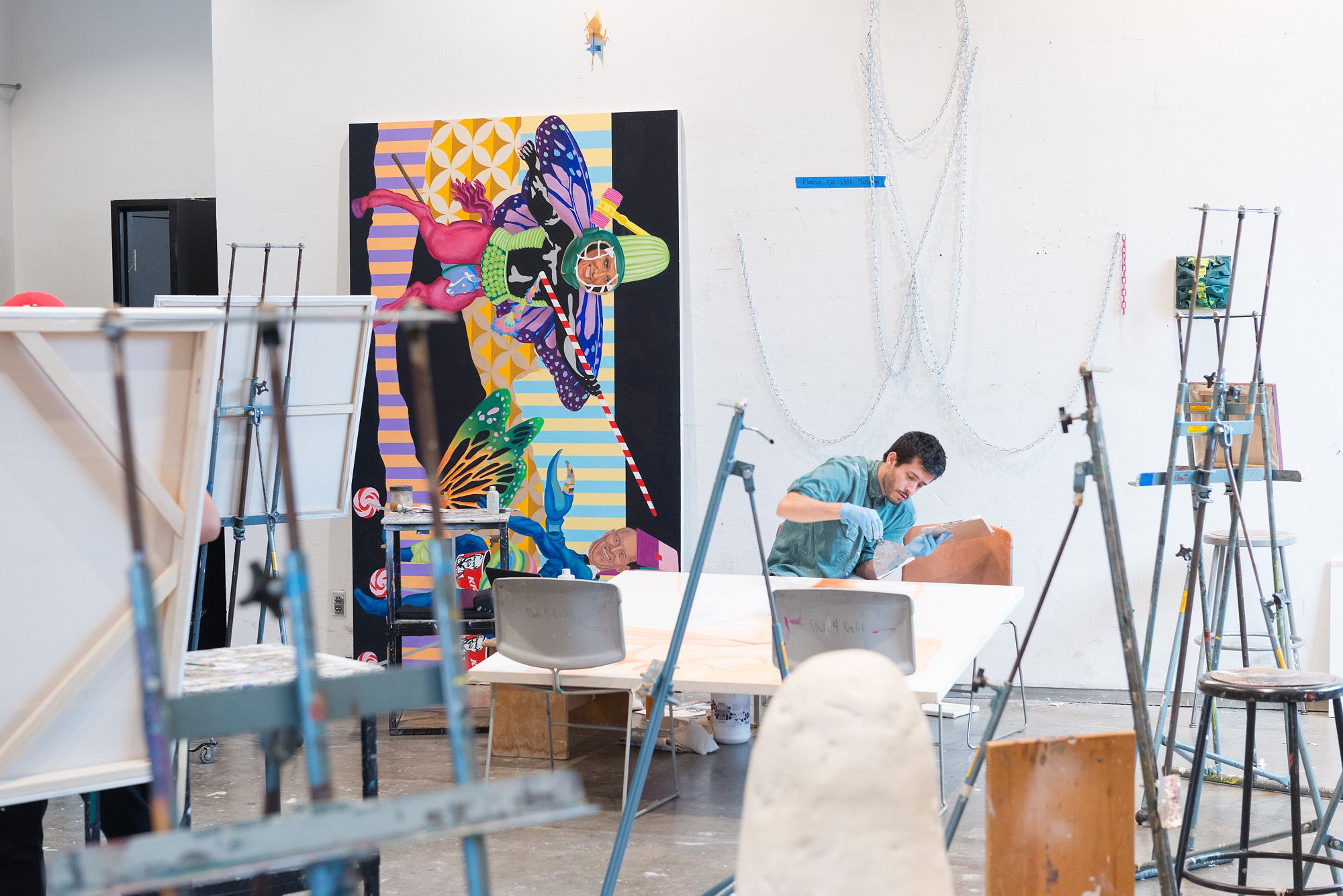 An art studio filled with easels, stools, and canvases. A large, colorful surreal painting stands against the back wall. To the right, an artist wearing gloves works at a table, surrounded by art supplies, with decorative chains hanging on the wall behind him.