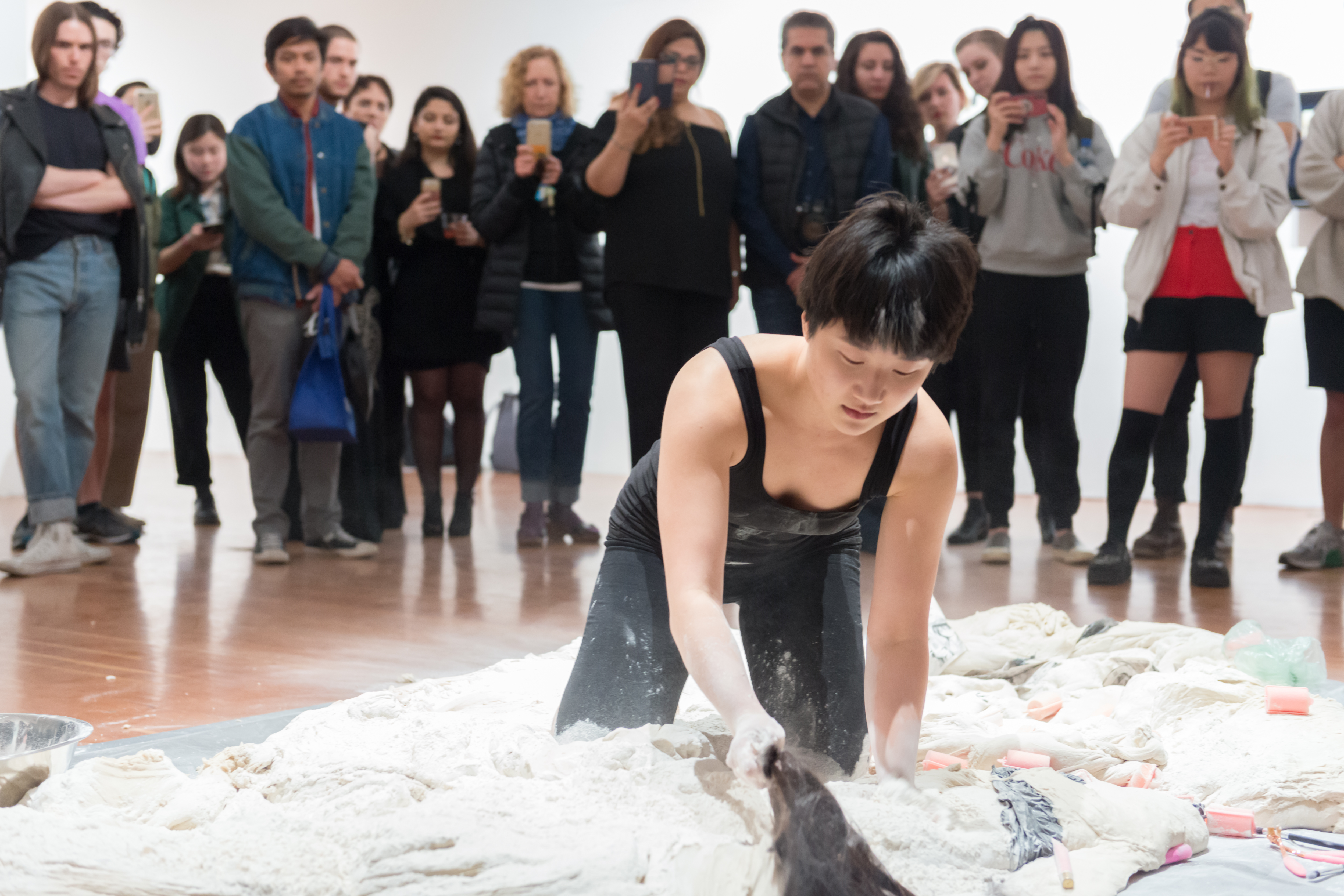 A performer kneels on the floor, dragging a long strand of hair through a pile of white powder and fabric, while a crowd of onlookers stands closely around, watching and recording the performance.