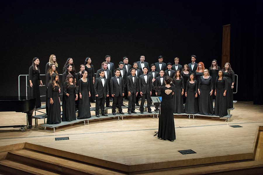 The UCI Choir performs on stage in Winifred Smith Hall, standing in formal attire and singing under the direction of a conductor.
