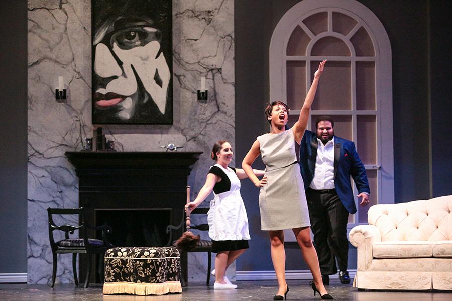 Opera singers perform on stage in a theatrical living-room set, singing and acting in costume during a staged production.