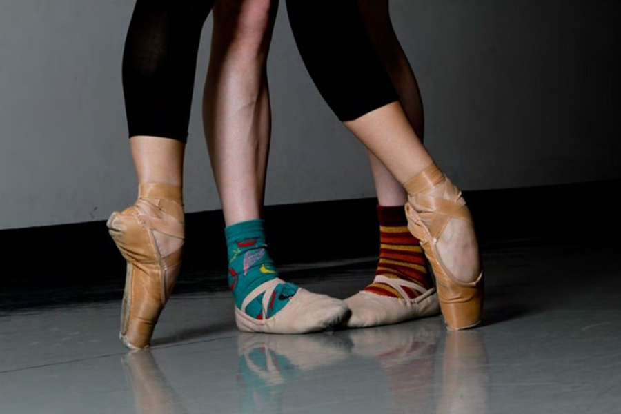 Close-up of two dancers’ legs in a studio, balancing on pointe shoes while wearing colorful mismatched socks.