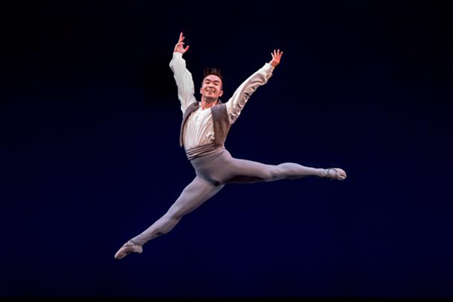 A male ballet dancer performs a grand jeté onstage, legs fully extended in midair with arms lifted and a joyful expression.