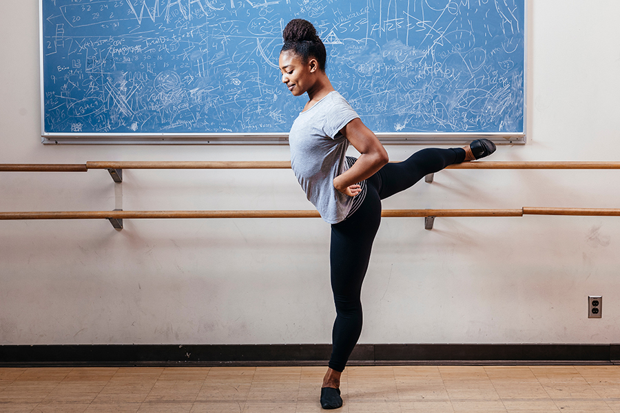 A dancer balances on one leg at the barre, extending the other leg behind her in an arabesque, with a chalkboard covered in drawings and notes in the background.