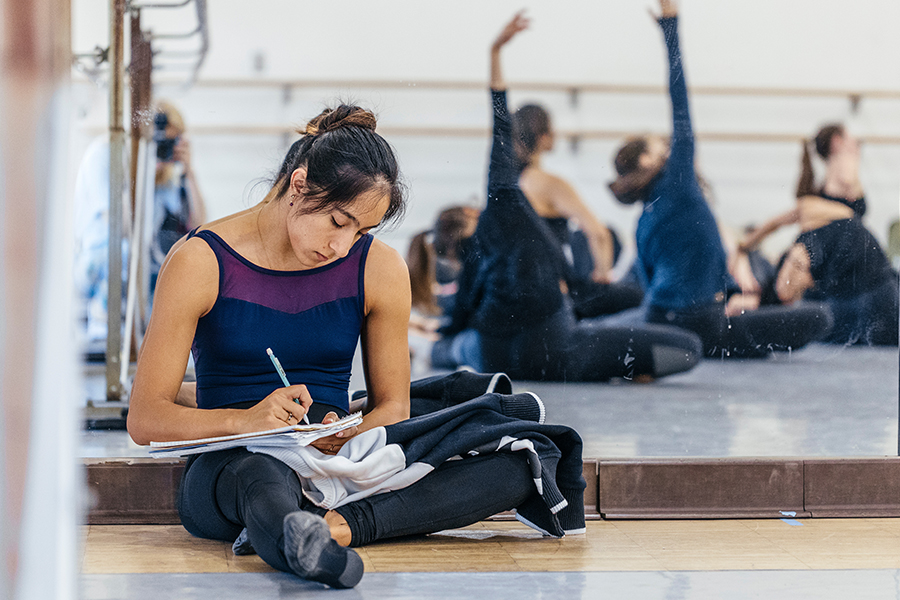 A dancer sits on the studio floor writing in a notebook, while other dancers stretch and rehearse in the mirrored background.
