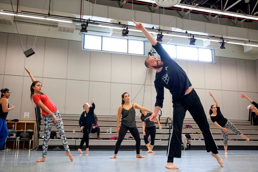A dance instructor leads students through a side-leaning movement in a large studio, with dancers spread out and mirroring his extended arm and body position.