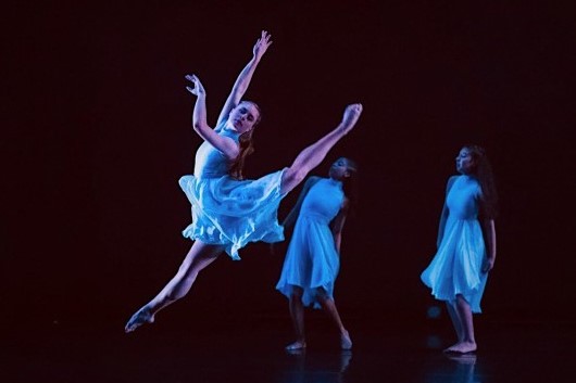 A dancer in a flowing blue dress leaps high onstage with arms and legs extended, while two other dancers in similar dresses watch from behind under dramatic lighting.