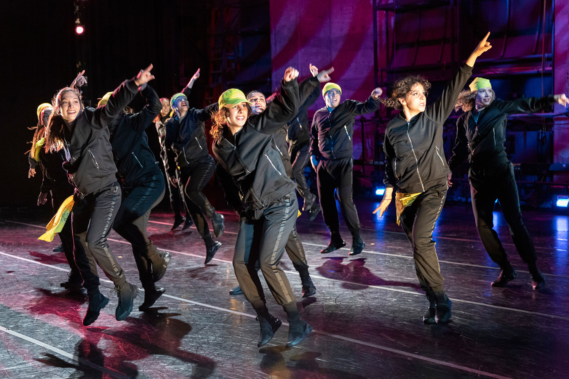 A group of dancers in matching black outfits and colorful headbands performs an energetic routine onstage, each pointing upward with lively expressions and movement.
