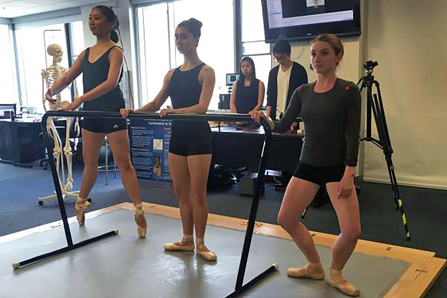 Three ballet students stand in a studio space holding a portable barre, practicing a position on pointe while instructors and equipment are visible in the background.