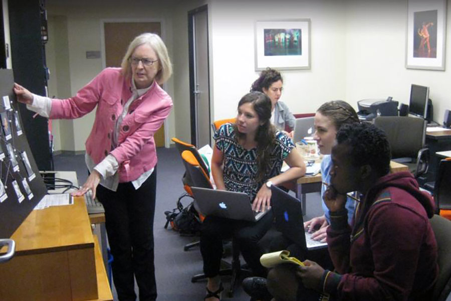 A faculty member gestures to a display board while a group of students with laptops listen and take notes in a classroom setting.