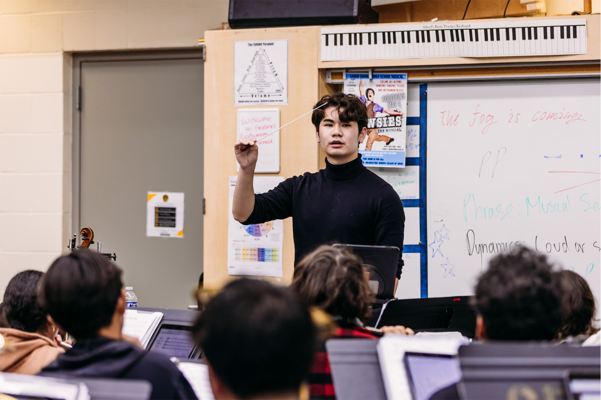 A student instructor leads a music class, holding a conductor’s baton while standing in front of a whiteboard, as seated students follow along with sheet music; the setting is a UCI Creative Connections classroom.