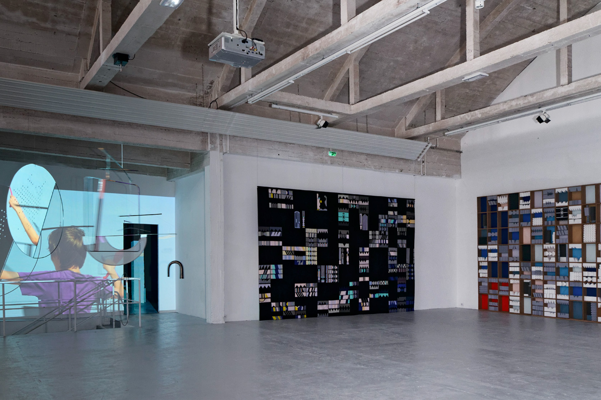 Interior of a contemporary art gallery with exposed concrete beams, featuring a projected video installation on glass and abstract geometric artworks mounted on white walls.
