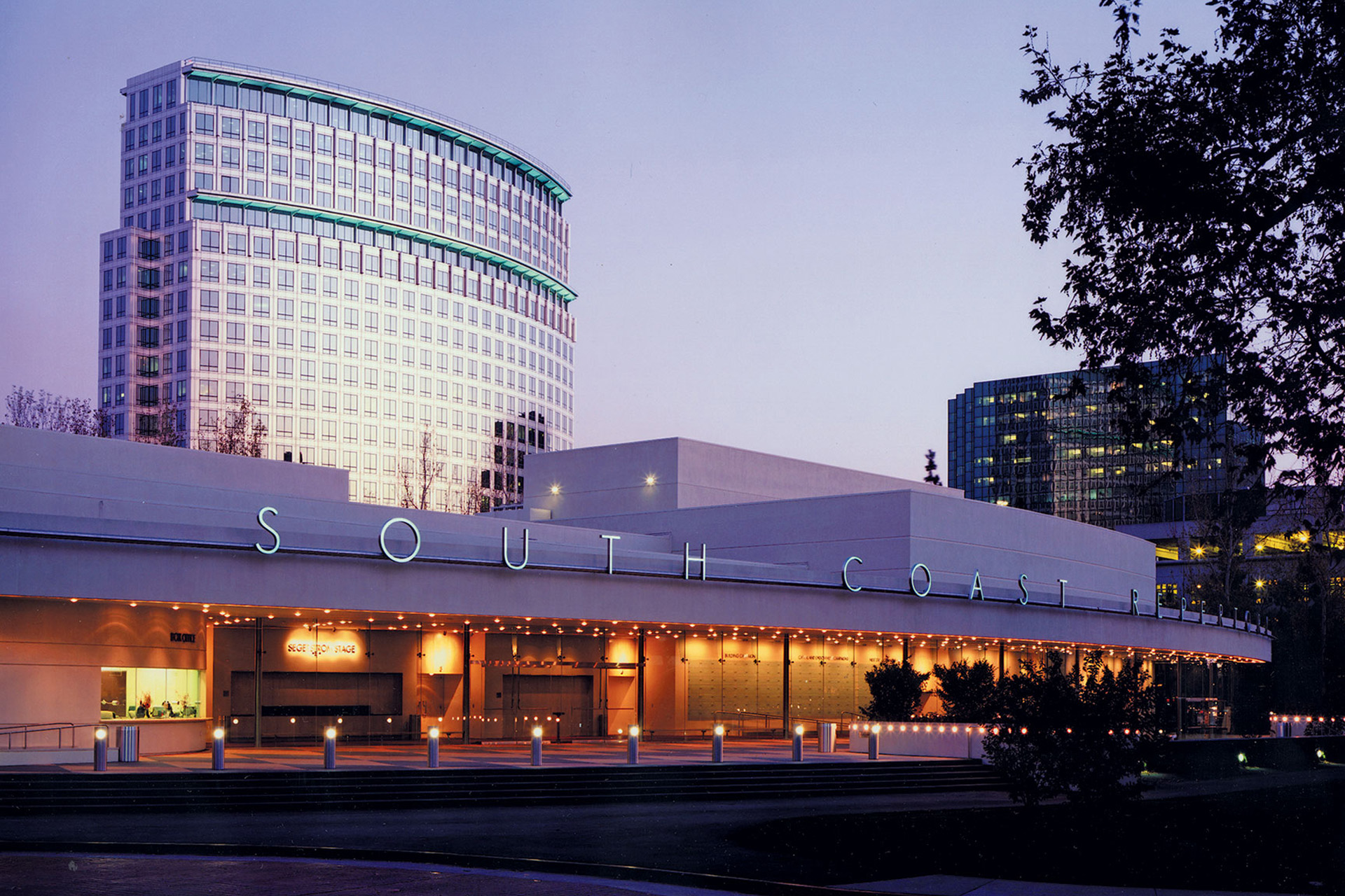 Evening view of the South Coast Repertory theater building with illuminated signage, warm exterior lights, and modern office towers in the background.