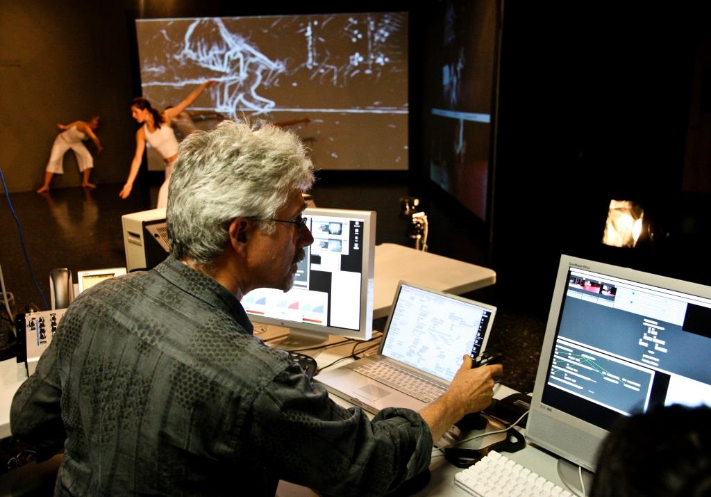 A person operates multiple computer screens and equipment in a darkened studio while dancers perform in the background against large projected visuals.