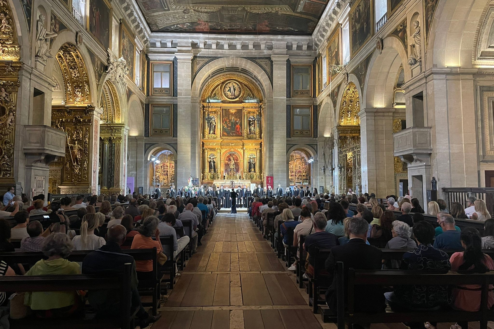 Interior of an ornate historic church with gilded arches and paintings, where a choir performs at the altar before a large seated audience.