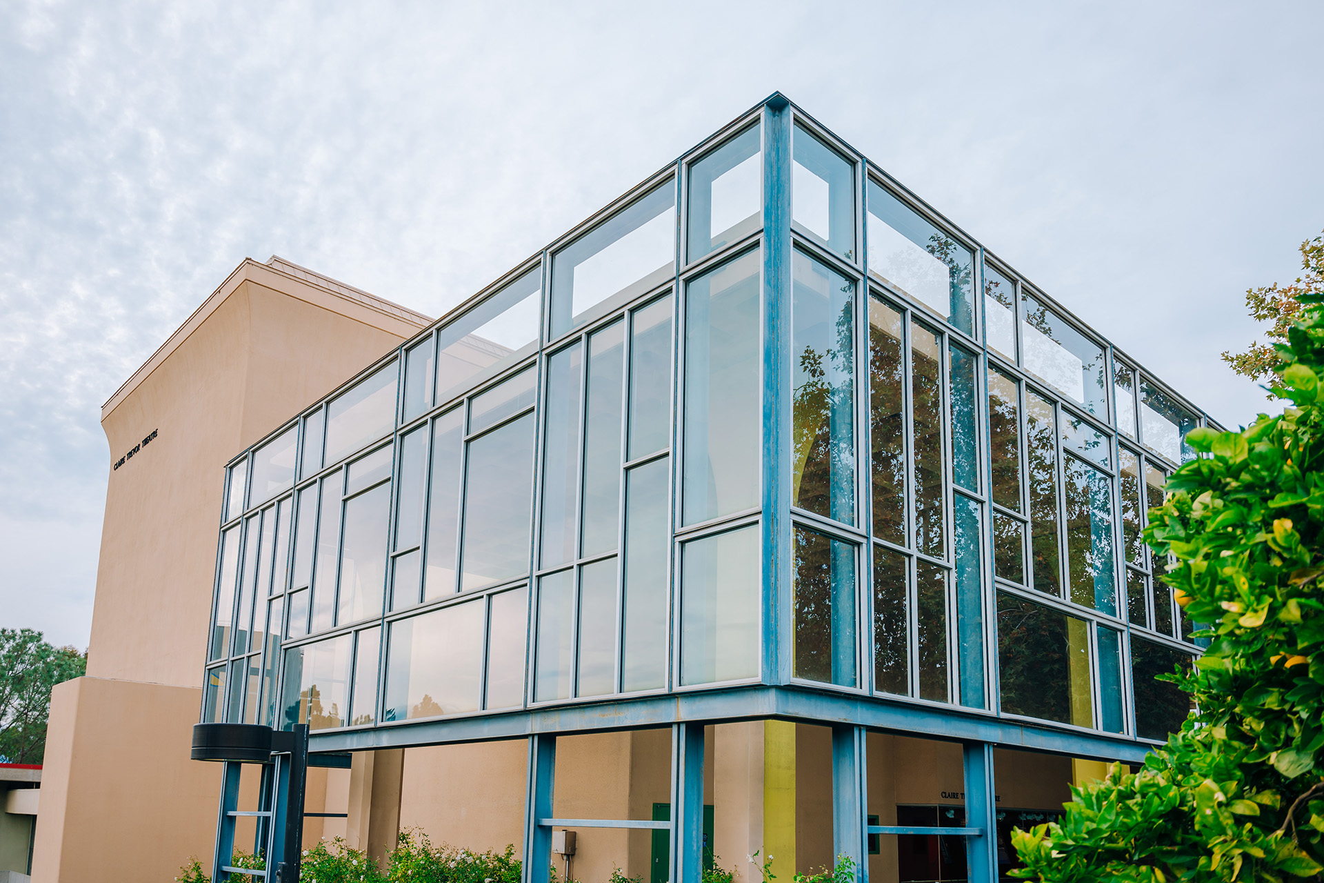 Exterior view of a modern glass-walled campus building (the Claire Trevor Theatre) with blue metal framing, reflecting trees and sky, surrounded by greenery.