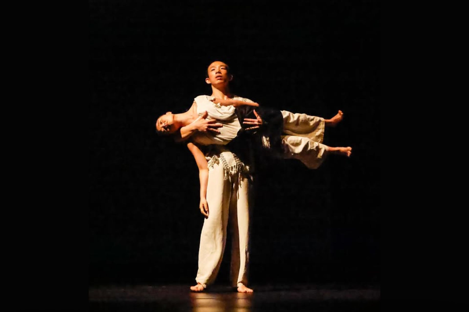 Two contemporary dancers perform a partnered lift on a dark stage, with one dancer standing and supporting another held horizontally across their torso under focused theatrical lighting.