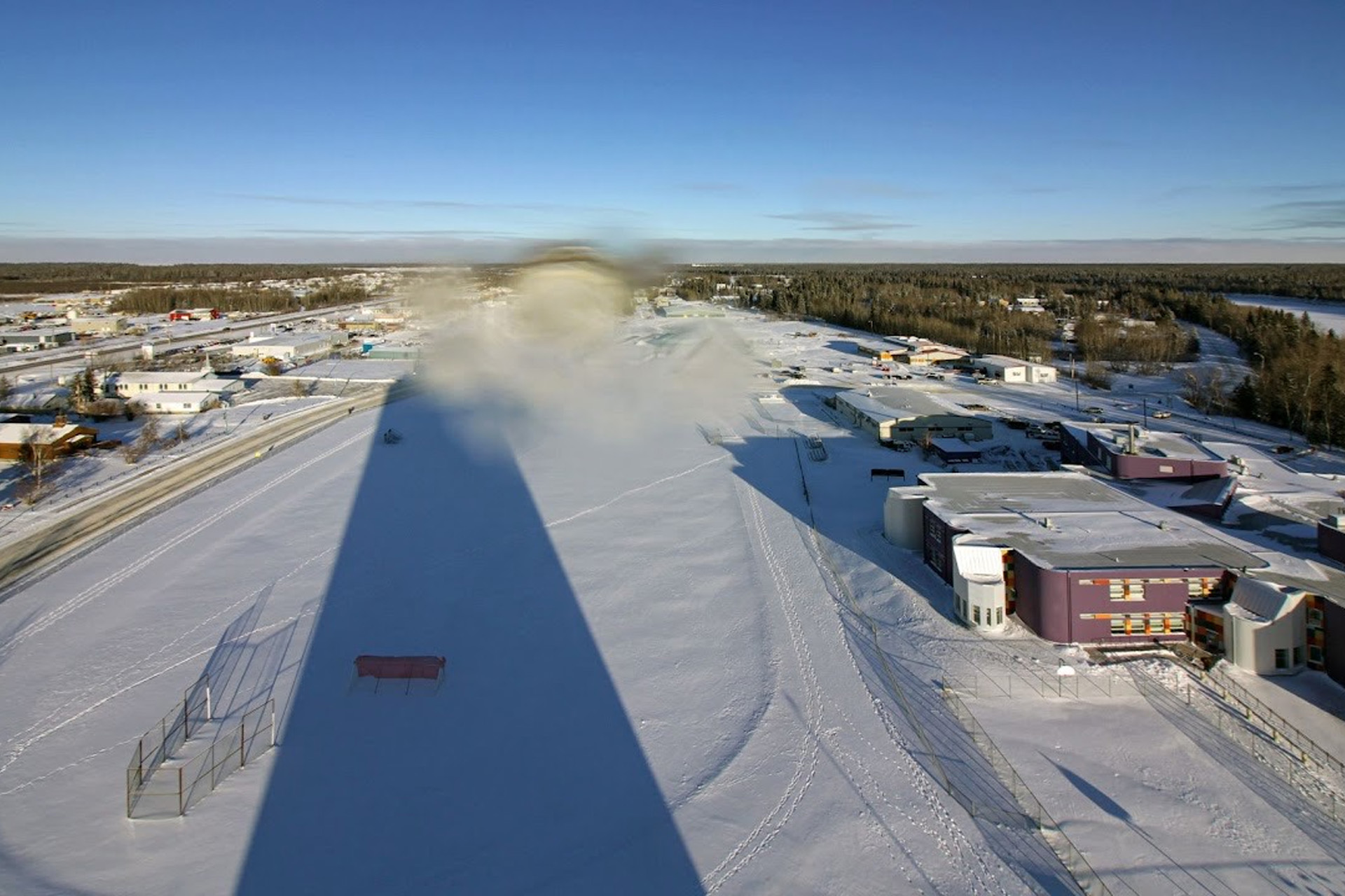 Aerial view of a snow-covered town under a clear blue sky, with buildings and roads visible and a tall structure casting a long shadow across the snowy ground.