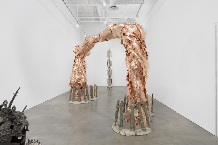 Large sculptural installation in a white gallery featuring arching, copper-toned forms that rise from the floor and curve inward, with textured vertical elements at their bases.