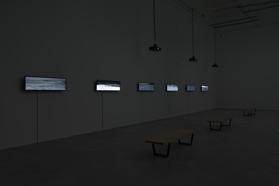 Row of small video screens mounted along a gallery wall, each displaying grayscale imagery, with benches positioned in front.