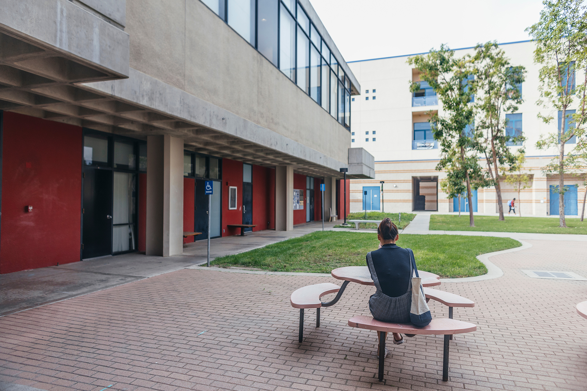 A student sits alone at a round outdoor table in a campus courtyard, surrounded by modern concrete buildings, grass, and walkways.