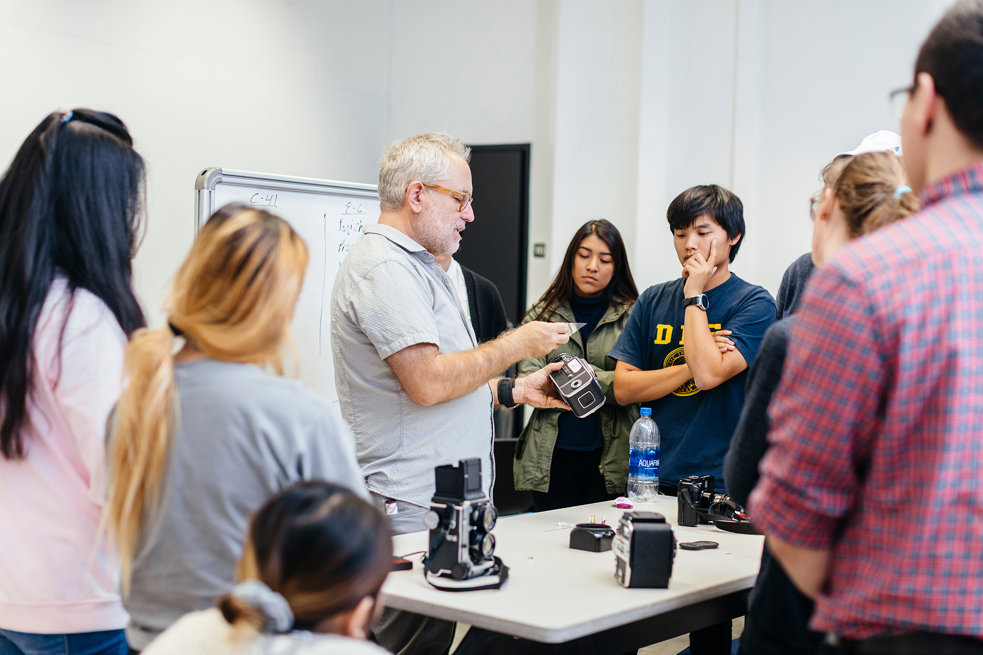 An instructor demonstrates a camera to a small group of students gathered around a table in a classroom.