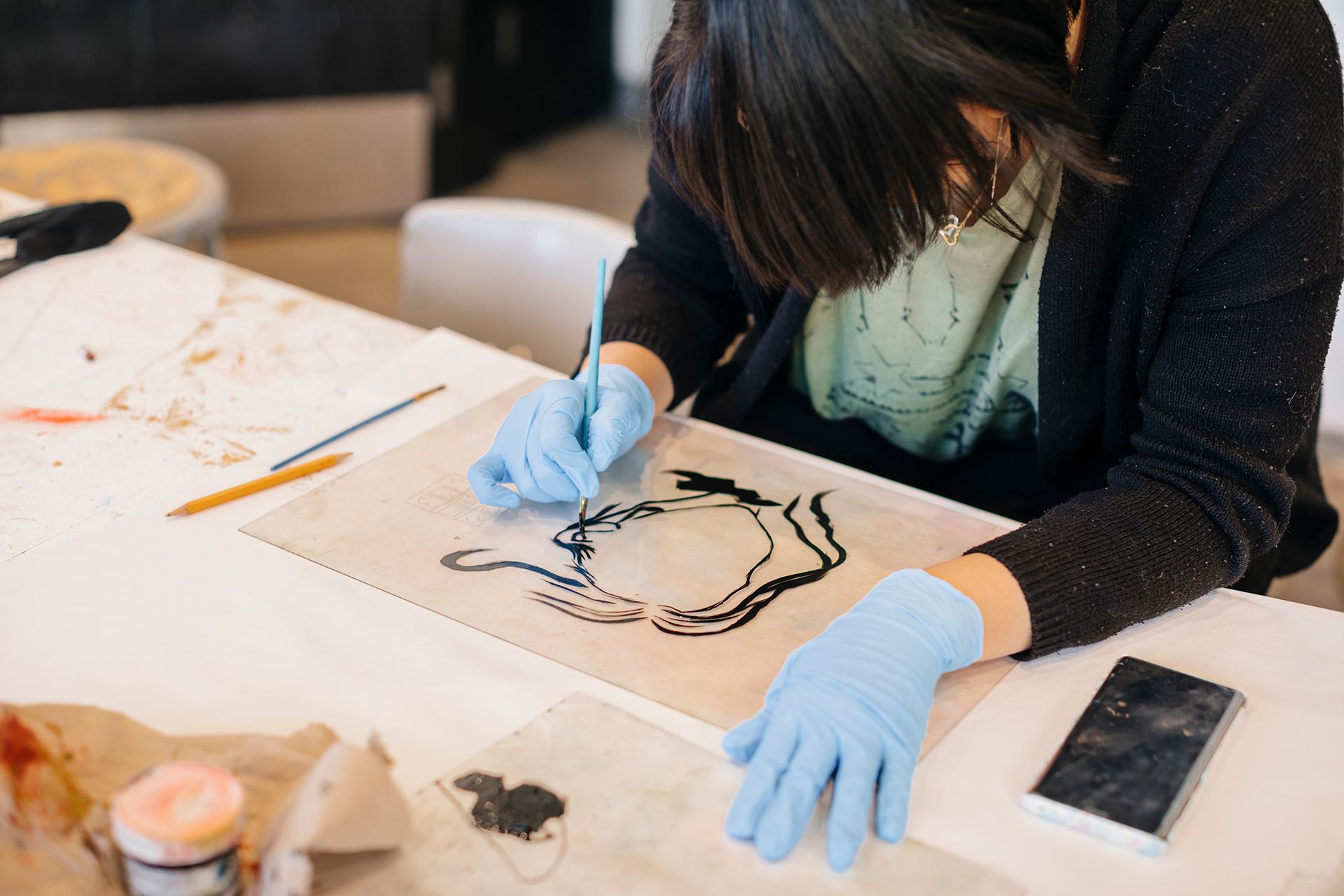 A student wearing blue gloves carefully applies black ink with a brush to a printmaking plate on a worktable.
