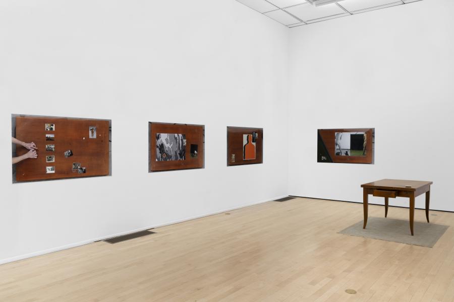 Gallery wall displaying mixed-media works mounted on wooden panels, with a small wooden table placed on a rug in the center of the room.