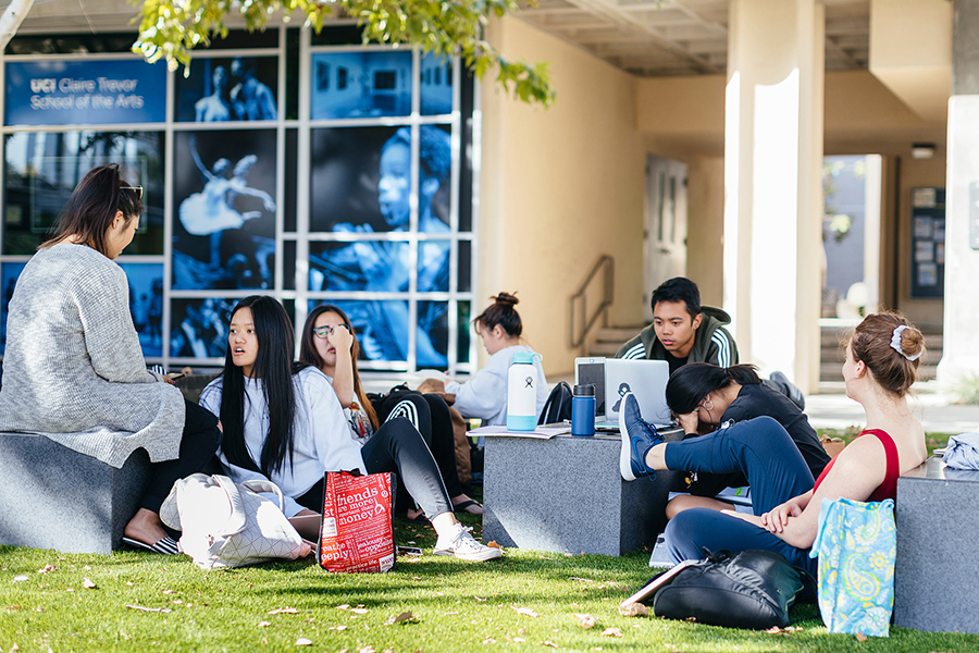 Students relax and study outdoors on campus, sitting on benches and grass with laptops and notebooks near the Claire Trevor School of the Arts building.