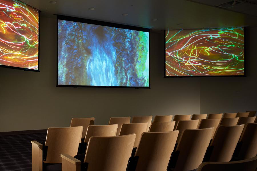 A small auditorium with tiered seating faces three large screens displaying abstract, colorful video imagery.