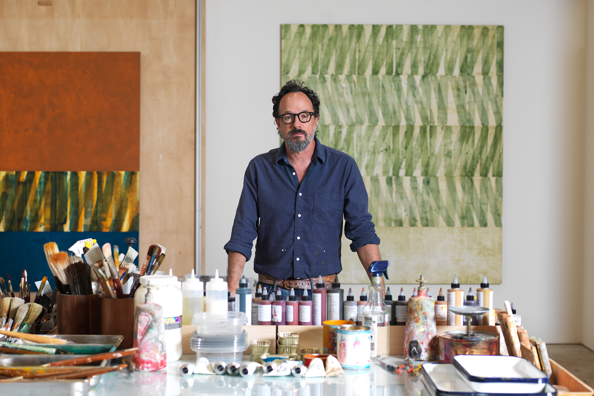  	Kevin Appel stands in his art studio behind a table of paints and brushes and in front of is paintings of green and rust. 