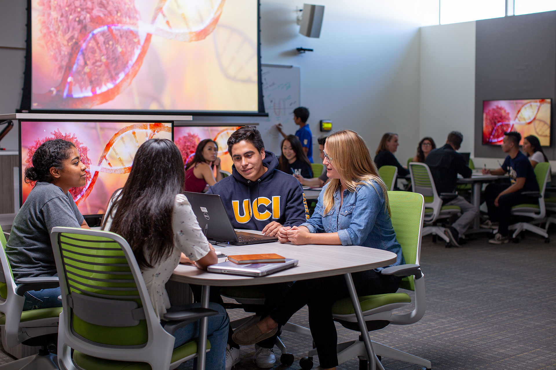 Students collaborate around a table in a modern classroom, reviewing work on a laptop as large screens display scientific imagery and other groups work in the background.