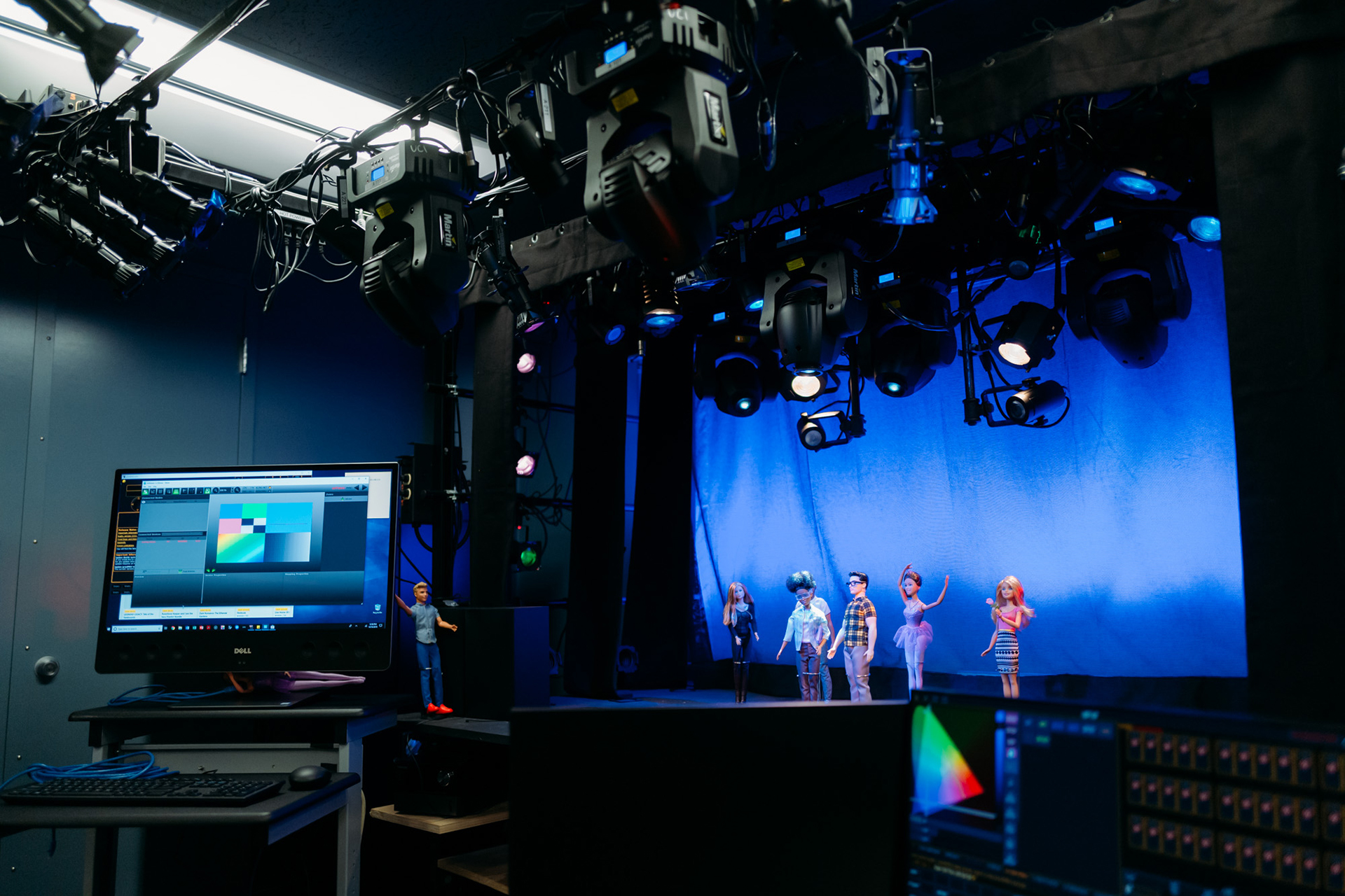 Lighting and projection design studio with theatrical lighting rigs overhead, computer monitors displaying color and cue controls, and scale figures staged against a blue-lit backdrop.