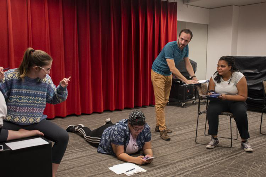 Students and an instructor collaborate during a directing rehearsal, with participants seated, standing, and working on the floor in front of a red curtain.
