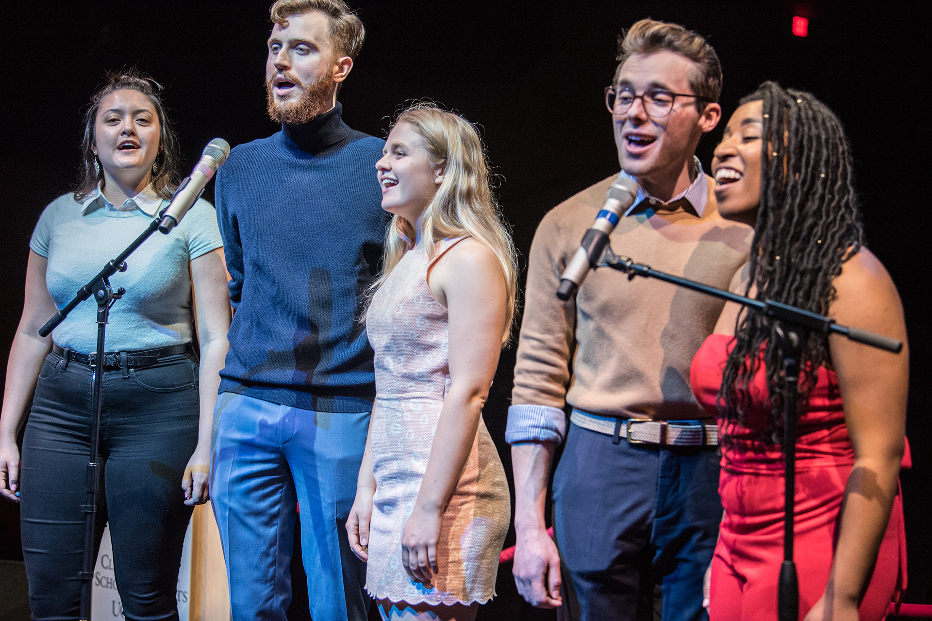 Music direction students sing together on stage at standing microphones, performing as an ensemble during a live vocal presentation.