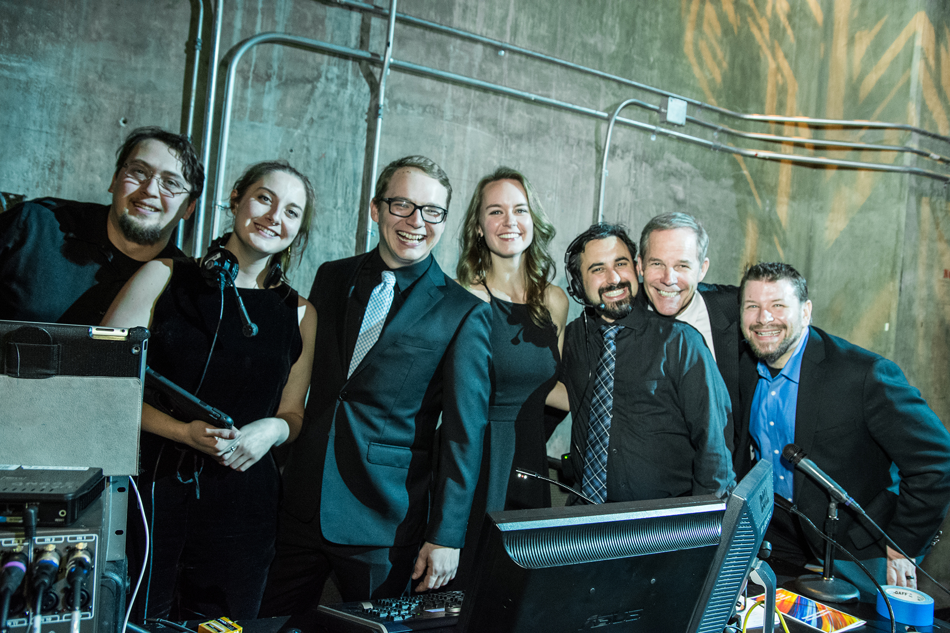 Stage management students and faculty pose together backstage, smiling beside sound and communications equipment in a theatre control area.