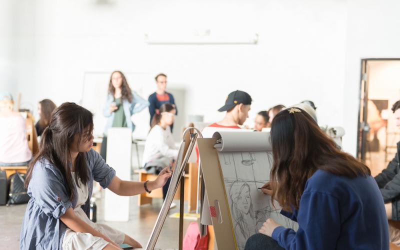 Students sketch from observation in a bright art studio, with one student drawing at a standing easel while others work and converse in the background.