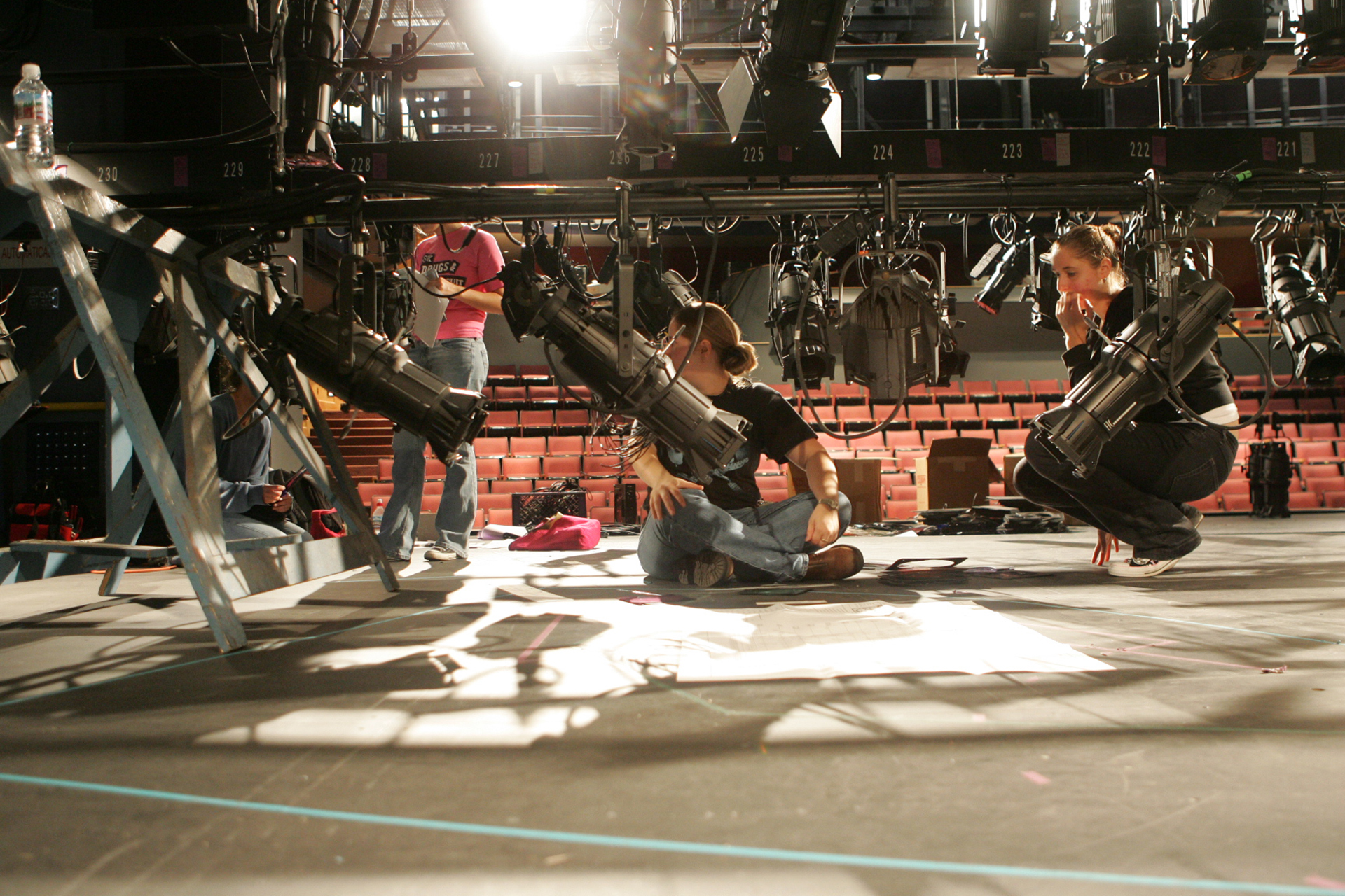 Students work beneath stage lighting instruments during a technical rehearsal, adjusting equipment and reviewing notes on a theater stage.