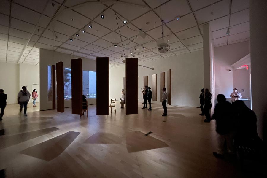 Visitors walk through a gallery installation composed of tall vertical panels and projected video, arranged across an open exhibition space.