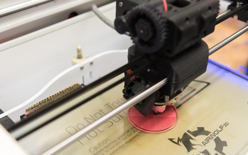 A 3D printer fabricates a small circular object in pink filament on a print bed.