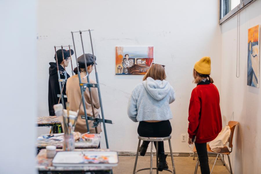 Students stand and sit in an art studio viewing and discussing a small painting mounted on a white wall, with easels and supplies nearby.