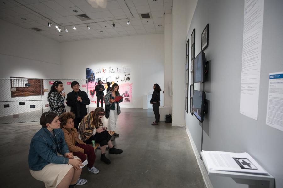 Visitors sit and stand in a gallery listening and viewing a wall-mounted video and artworks during an exhibition.