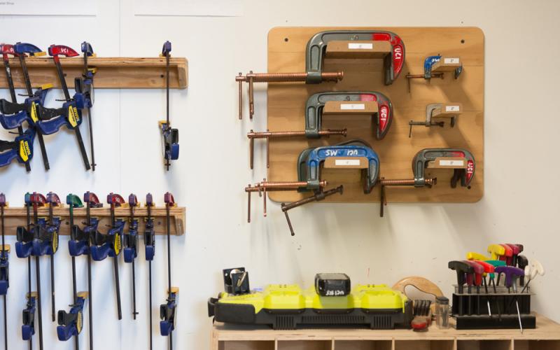 A workshop wall displays neatly organized clamps, hand tools, and woodworking equipment.