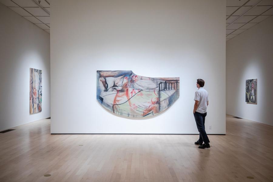 A person stands in a museum gallery viewing a large, curved drawing mounted on a white wall, with additional works displayed nearby.