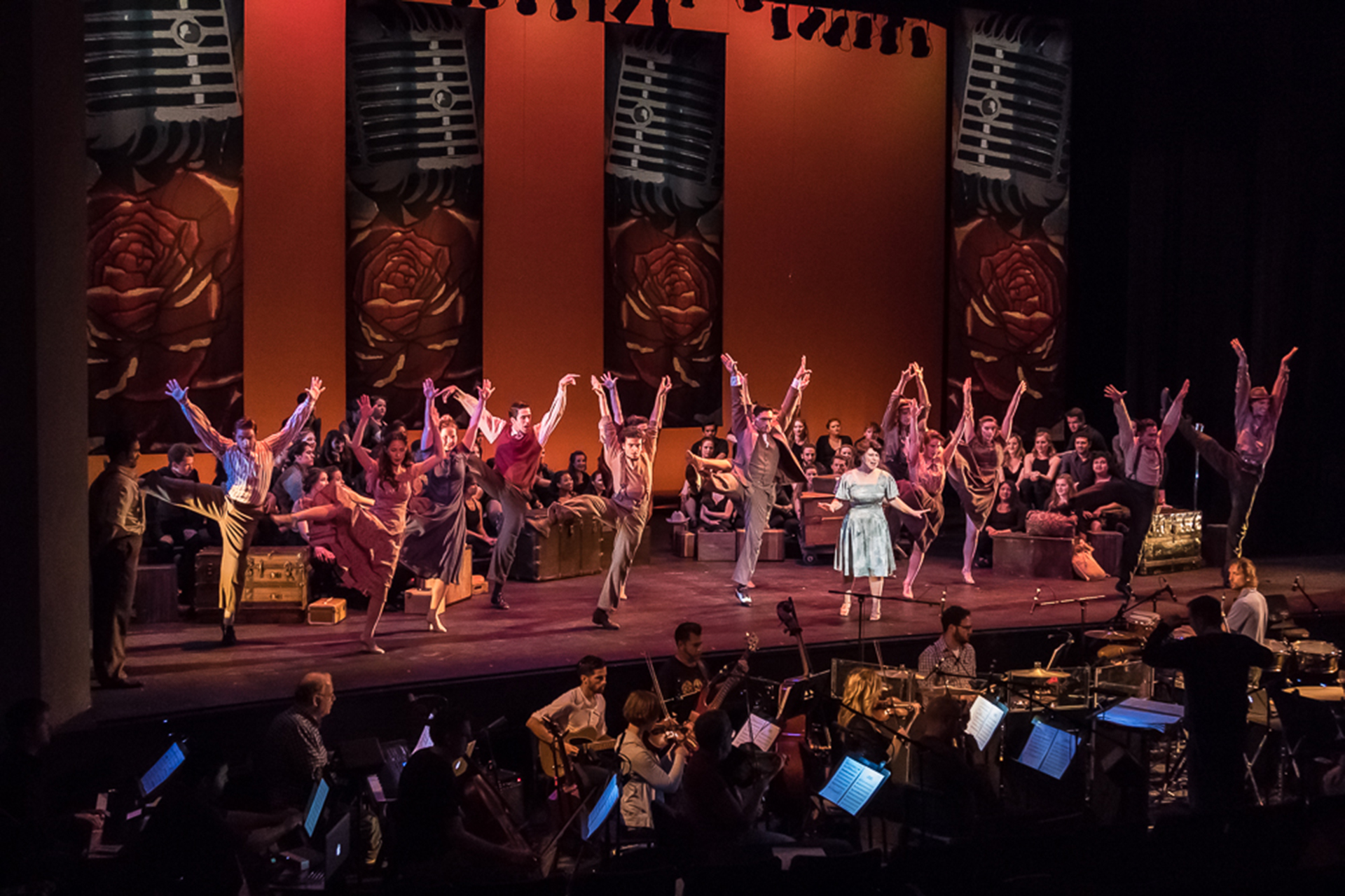 Large ensemble of music theatre students perform a choreographed number onstage with a live orchestra visible in the pit below.