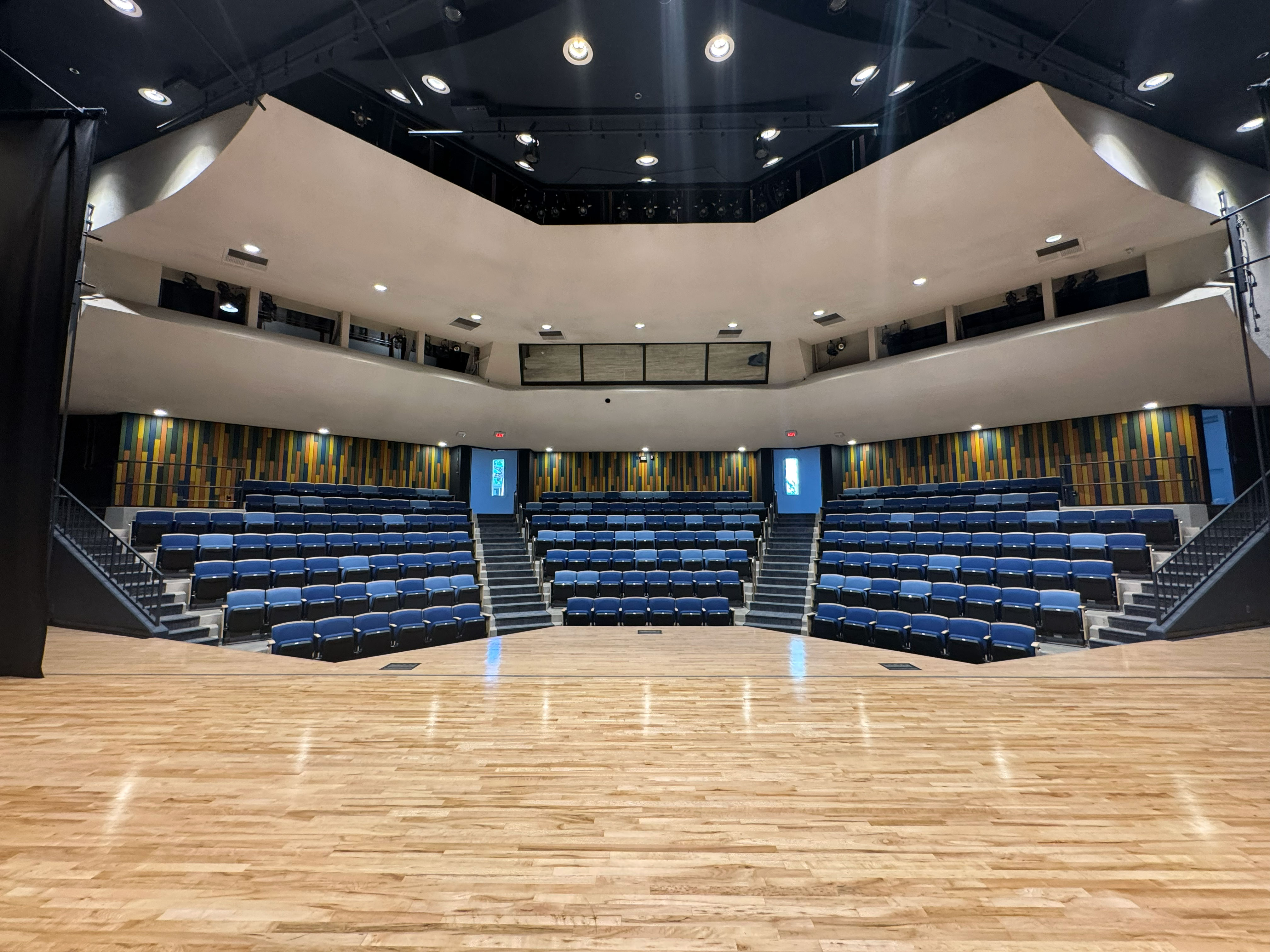 View from the stage of a modern theater with a polished wood floor and tiered seating filled with blue chairs, surrounded by curved balconies, colorful acoustic wall panels and overhead stage lighting.