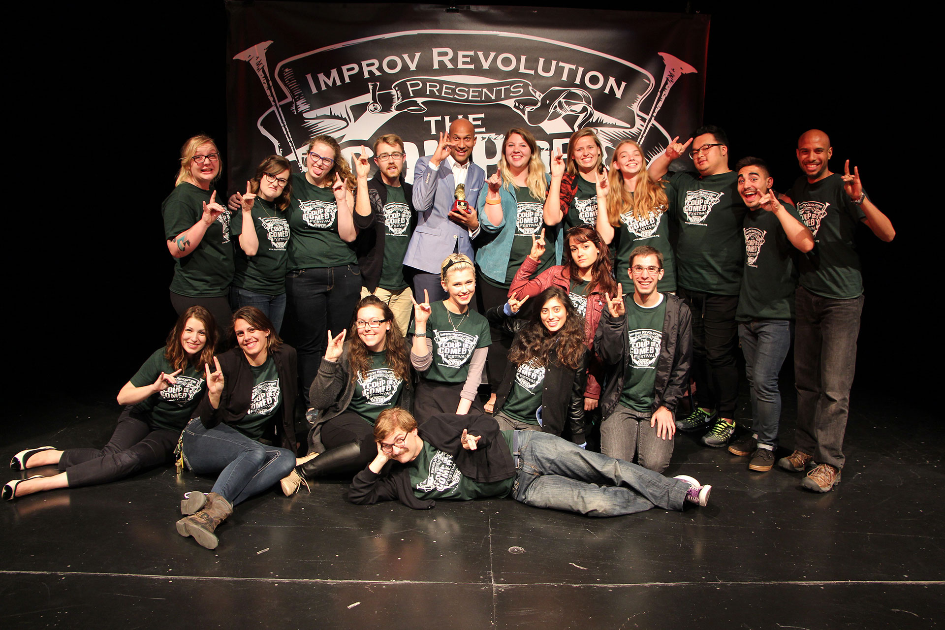 Improv comedy group in matching festival T-shirts pose on stage with a man in a blue suit holding an award beneath an “Improv Revolution Presents” banner.