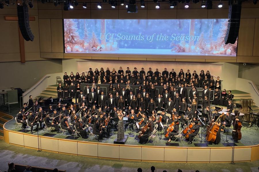 Wide shot of choir and orchestra on a concert-hall stage with a conductor at center; a large screen above reads "UCI Sounds of the Season" over a snowy winter scene.